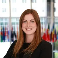 A woman with long brown hair and a friendly smile is standing against a backdrop of flags from various countries, dressed in a black blazer.