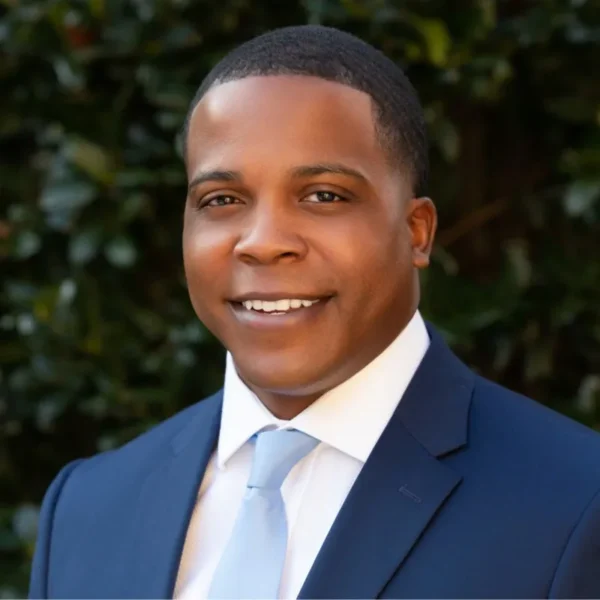 A smiling man wearing a dark suit with a light blue tie, standing outdoors against a green foliage background.