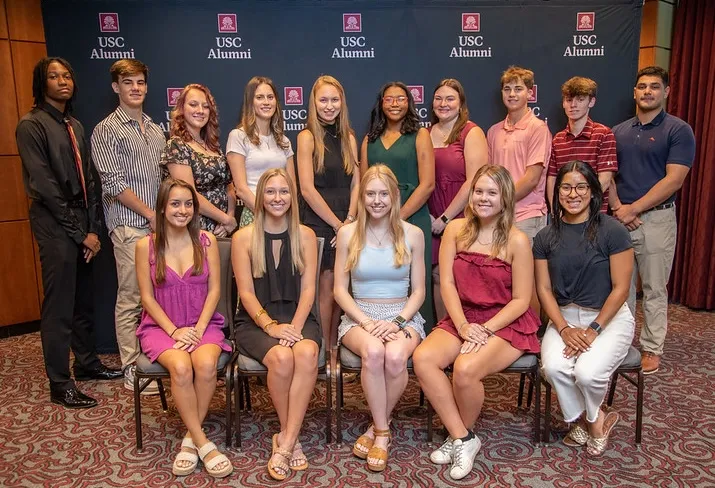 A group photo of sixteen individuals, arranged in two rows. The front row consists of eight young women seated, while the back row features eight individuals, including both male and female students, some standing and some slightly turned. They are posed in a formal setting with a backdrop displaying 'USC Alumni'. Everyone is dressed in various styles of casual and semi-formal clothing.