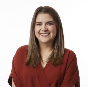 A young woman with long brown hair is smiling, wearing a rust-colored blouse against a plain white background.