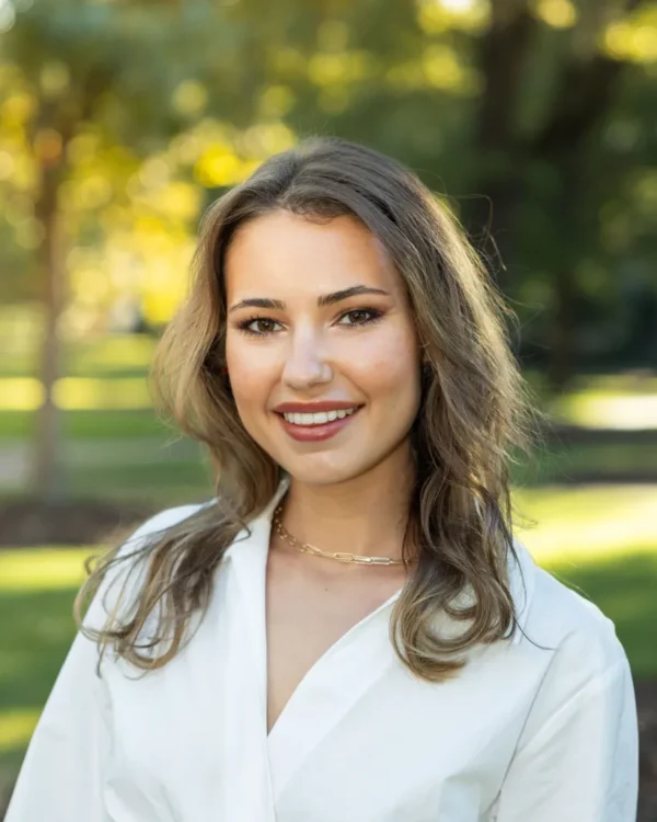 A young woman with wavy brown hair is smiling at the camera, wearing a white blouse and a simple necklace. She stands in a park with soft greenery and sunlit bokeh in the background.