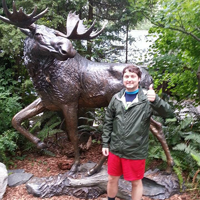 A man in a green jacket and red shorts stands beside a bronze statue of a moose, giving a thumbs up, surrounded by lush greenery.