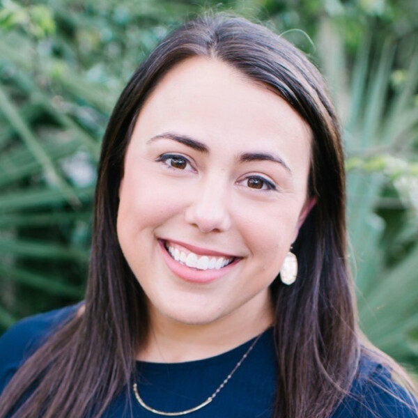 A woman with long dark hair and a friendly smile is wearing a dark blue top and a delicate necklace. In the background, there are green plants, suggesting an outdoor setting.