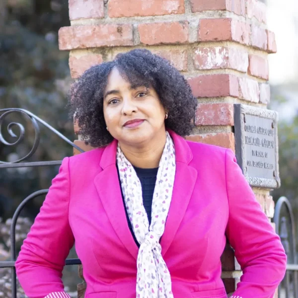 A woman with curly hair, wearing a bright pink blazer and a patterned scarf, leans against a brick column with a plaque. She appears to be in a garden setting, looking directly at the camera with a confident expression.