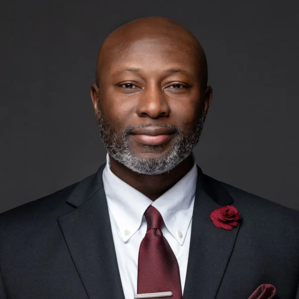 A well-dressed man with a bald head and a greying beard, wearing a black suit, white shirt, and a maroon tie. He has a red rose pinned to his lapel and is posed against a dark background, exuding a confident demeanor.