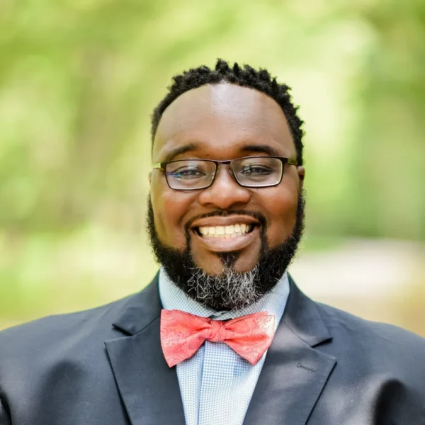 A man with glasses smiles broadly, wearing a navy suit and a pink bow tie, with a light blue checkered shirt underneath, set against a blurred green natural background.