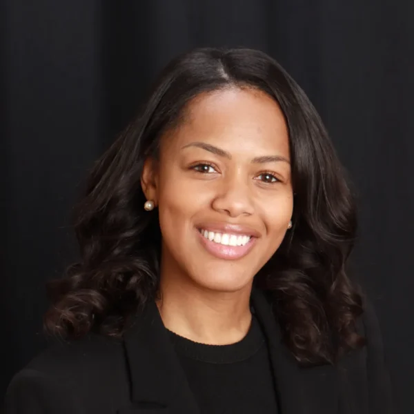 A woman with curly black hair smiles warmly at the camera, wearing a black blazer over a black top, with a dark background behind her.