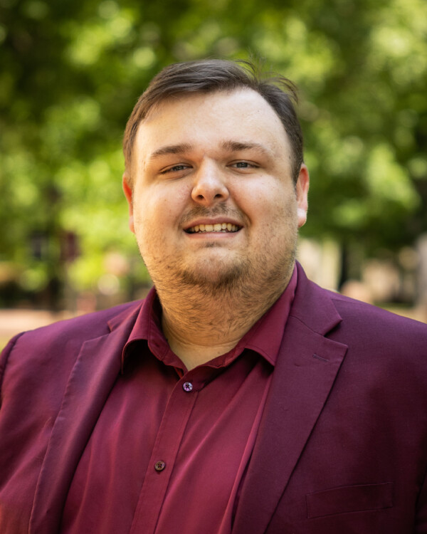 A smiling man with light brown hair and a short beard, wearing a burgundy blazer over a matching shirt, stands outdoors in front of a blurred green background.