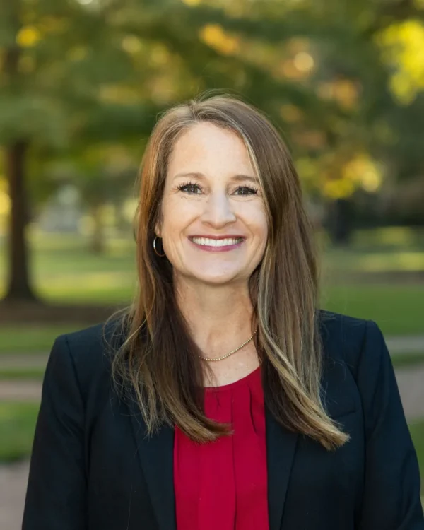 A smiling woman with long, brown hair stands outdoors in a park setting, wearing a black blazer over a red blouse. The background features blurred trees and green grass, suggesting a sunny day.