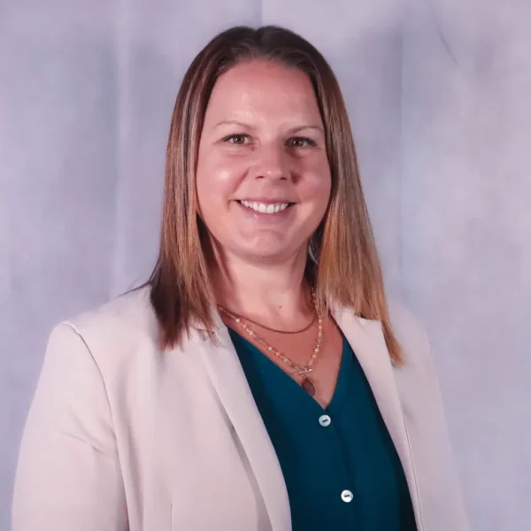A woman with long, straight hair smiles at the camera, wearing a light-colored blazer over a green blouse. She has layered necklaces and a neutral background.