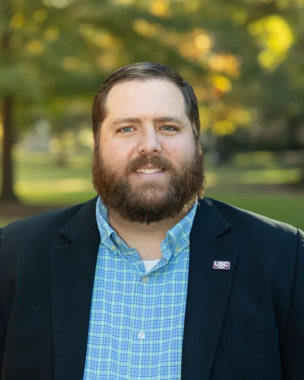 A man with a beard wearing a dark blazer over a light blue and green plaid shirt, standing outdoors with blurred trees in the background. He has a pin on his blazer featuring the letters 'USC'.