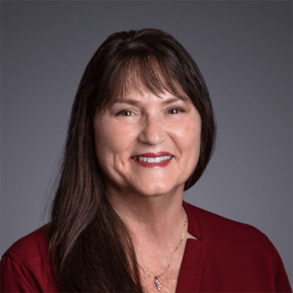 A woman with long dark hair and bangs smiles in front of a gray background, wearing a burgundy top and a necklace.