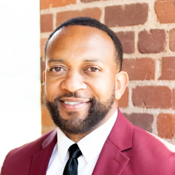 A man with a beard wearing a burgundy suit jacket and a white shirt, smiling against a backdrop of exposed brick.