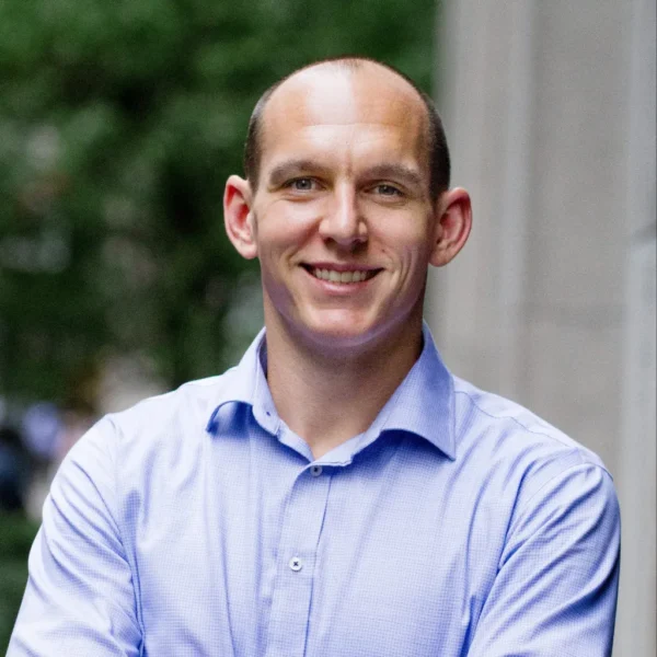 A man with a neatly shaved head, smiling while standing outdoors with his arms crossed, wearing a light blue button-up shirt. The background features greenery and a blurred cityscape.