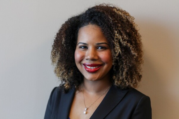 A woman with curly, shoulder-length hair featuring highlights, wearing a black blazer and a necklace, smiling against a neutral background.