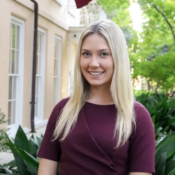 A young woman with long, straight blonde hair smiles while standing in a garden area, wearing a burgundy dress. The background features green foliage and a building with large windows.