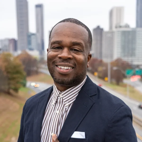 A man smiling confidently while wearing a navy blazer over a striped shirt, standing against a backdrop of a city skyline. The blurred view of a highway is visible in the foreground, suggesting an urban setting.