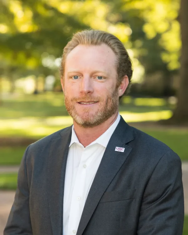 A man with a light beard, wearing a dark suit and white shirt, stands outdoors with a blurred green background. He has a pin labeled 'USC' on his left lapel.