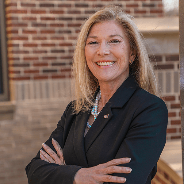 A woman with long blonde hair smiles confidently with her arms crossed, wearing a black blazer and a necklace with pearls, standing against a backdrop of brick and stone architecture.