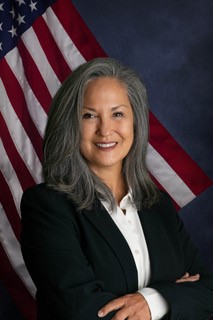 A woman with long gray hair smiling confidently while wearing a black blazer and white shirt, standing in front of an American flag backdrop.
