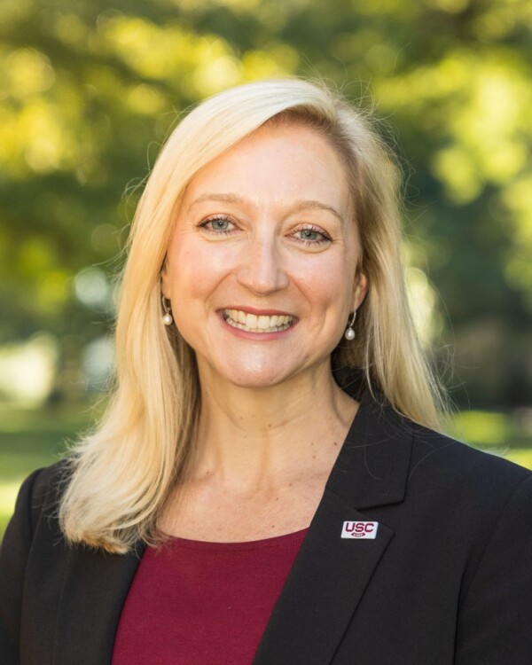 A woman with long blonde hair smiling while wearing a black blazer over a maroon top, with a USC pin on her blazer, set against a blurred green park background.