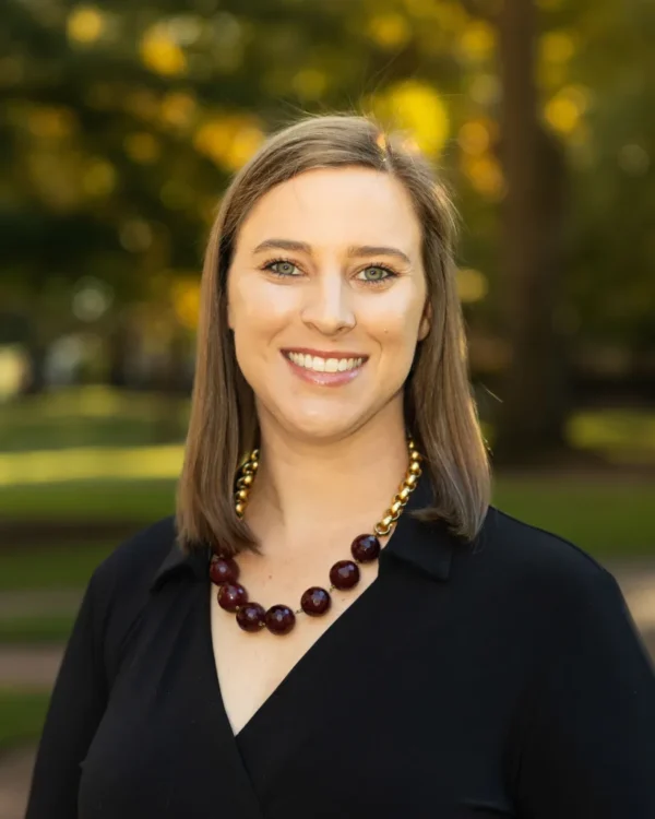 A woman with shoulder-length brown hair and green eyes smiles at the camera, wearing a black blouse and a chunky necklace featuring large maroon beads. The background is a softly blurred park scene with golden autumn foliage.