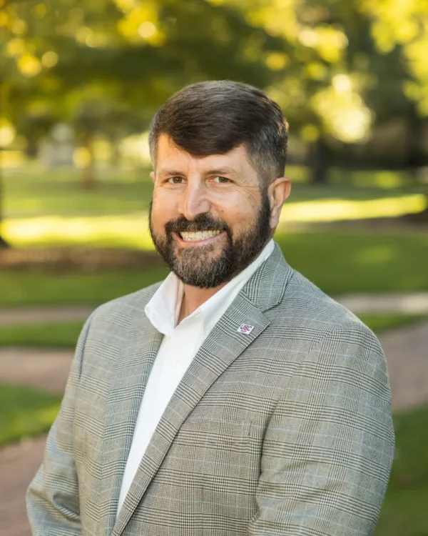 A man with a beard and short hair, wearing a light gray checkered blazer over a white shirt, smiles while standing in a park with greenery in the background.