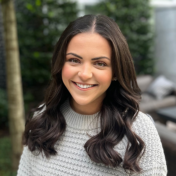 A young woman with long, wavy brown hair smiles warmly at the camera while wearing a cozy gray sweater. She appears in a lush outdoor setting with green foliage in the background.