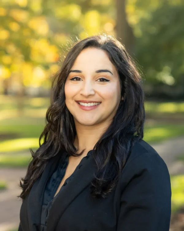 A woman with long dark hair, wearing a black blazer, smiles warmly in a sunlit park setting with blurred green and golden foliage in the background.
