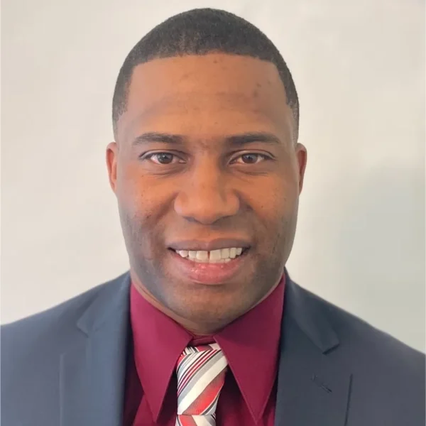 A man smiling at the camera, wearing a dark suit jacket and a maroon shirt, along with a striped tie that has red, white, and black colors, against a light gray background.