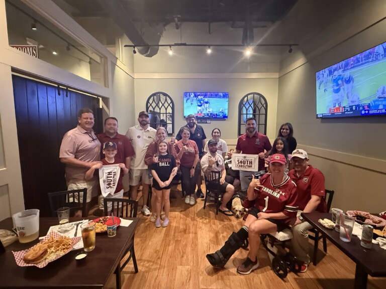 A group of people, wearing South Carolina Gamecocks apparel, gathers in a restaurant to celebrate, with banners and signs displaying pride for the team. Several TV screens show sports games, and a table displays food and drinks.