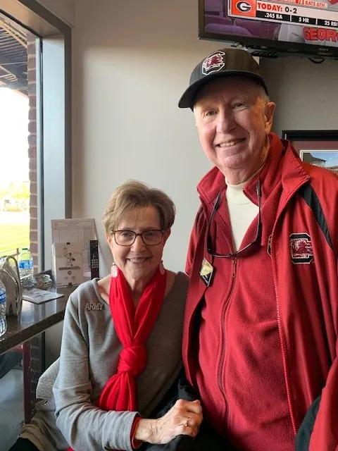 A smiling woman with short, wavy hair wearing glasses and a gray jacket with a red scarf stands next to an older man with short hair, wearing a red jacket and a black cap. They are in a bright setting with a large window, and there's a scoreboard visible in the background.