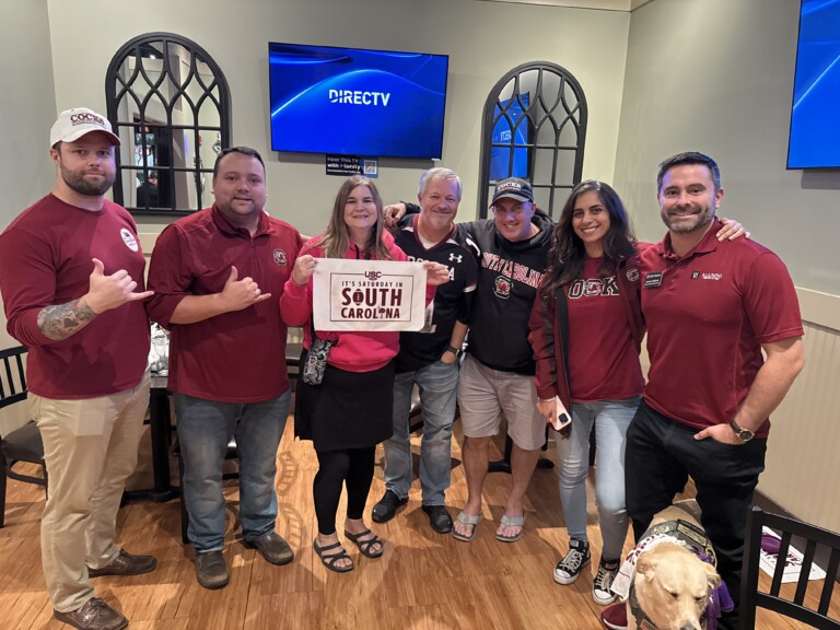 A group of seven people posing together in a casual setting, all wearing clothing in garnet and black, representing the University of South Carolina. One person holds a sign that reads "It's Saturday in South Carolina." A yellow dog is sitting nearby.