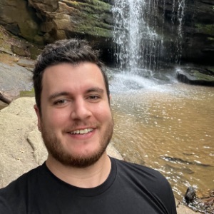 A person with short, dark hair and a beard smiles at the camera while standing near a waterfall, with rock formations and a pool of water in the background.