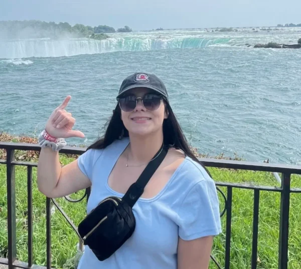 A smiling woman with long dark hair wearing a light blue shirt, sunglasses, and a black cap, standing in front of Niagara Falls and showing a hand gesture, with water cascading in the background and green grass at her feet.