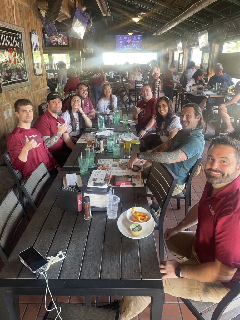 A group of eight people sitting at a table in a restaurant, wearing matching maroon shirts. They are smiling and posing for the camera, surrounded by drinks, menus, and food on the table, with a lively restaurant atmosphere visible in the background.