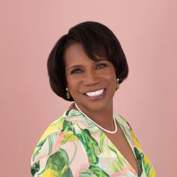 A smiling woman with short, dark hair stands against a pale pink background, wearing a colorful floral print blouse with green and pink hues, complemented by pearl and green earrings.