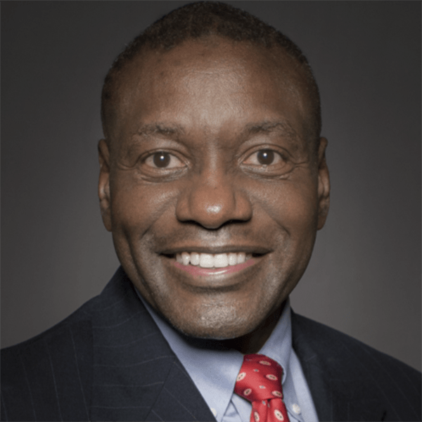 A man smiling confidently, wearing a dark suit with stripes and a red tie, against a neutral gray background.