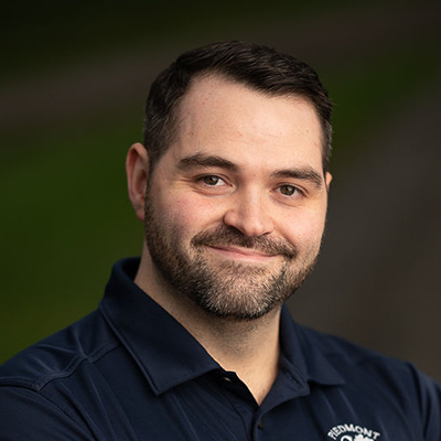 A man with a beard smiles confidently, wearing a dark blue polo shirt with a logo, set against a blurred green background.