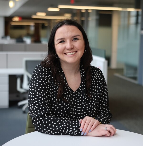 A woman with long brown hair, wearing a black and white polka-dotted shirt, smiles while sitting at a round table in a modern office environment with cubicles in the background.