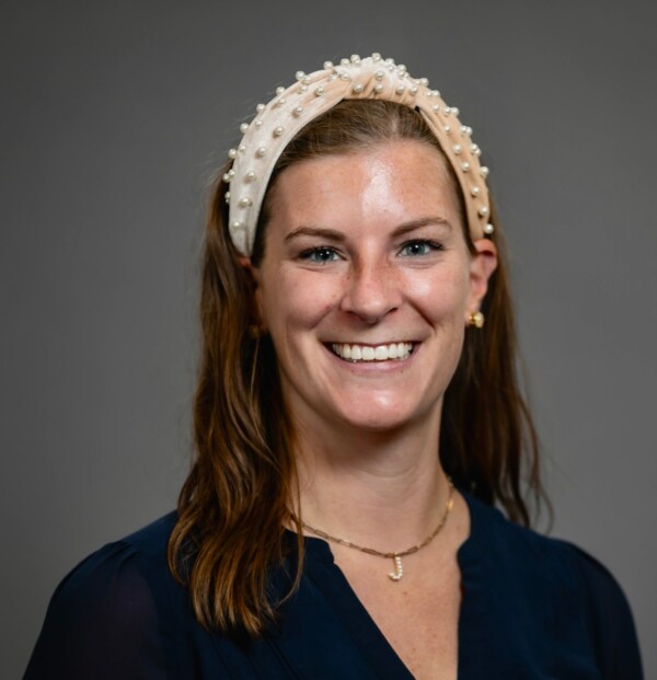A smiling woman with long, wavy brown hair wearing a decorative headband with pearls, a dark blue blouse, and a necklace featuring a 'J' pendant, set against a gray background.