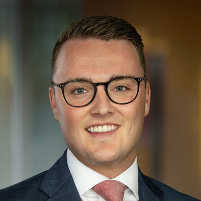 A young man with short, light brown hair and glasses is smiling at the camera, wearing a dark blue suit and a light pink tie, with a blurred indoor office environment in the background.