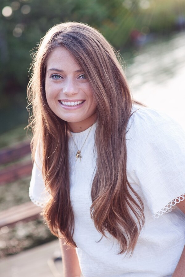 A woman with long, wavy brown hair and bright blue eyes smiles at the camera, wearing a white blouse with lace trim. She is outdoors, with a blurred background of green trees and water, basking in soft sunlight.