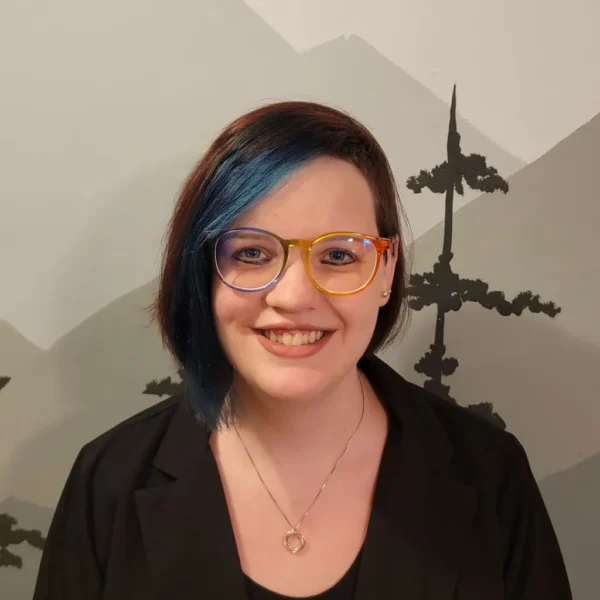 A woman with short, partially blue hair wearing colorful glasses and a black blazer smiles in front of a mountain-themed mural.