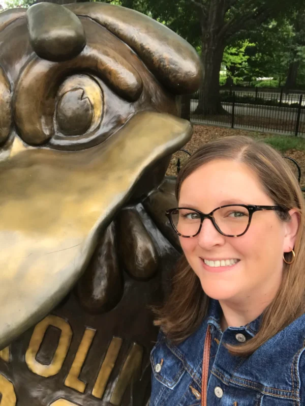 A woman with glasses smiles for a selfie beside a large bronze sculpture of a bird's head, featuring detailed textures, with trees and a fence visible in the background.