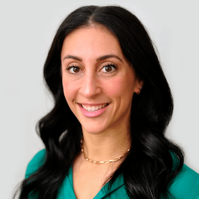 A smiling woman with long, dark hair wearing a teal blouse and a gold chain necklace against a light gray background.