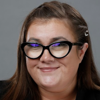 A woman with long brown hair wearing large black cat-eye glasses and a black blazer, smiling against a gray background.