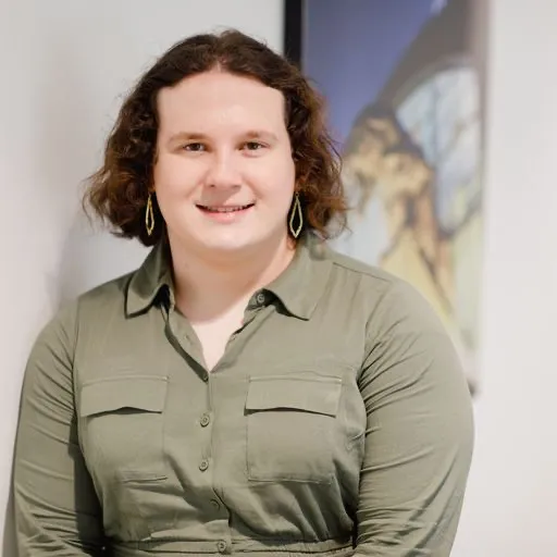 A smiling person with curly hair, wearing a green button-up shirt and large earrings, stands against a light background with a colorful artwork partially visible behind them.