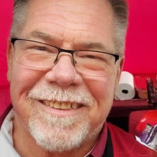 A smiling older man with glasses and a beard, wearing a red and gray shirt, against a background with a bright red color and some indistinct objects behind him.