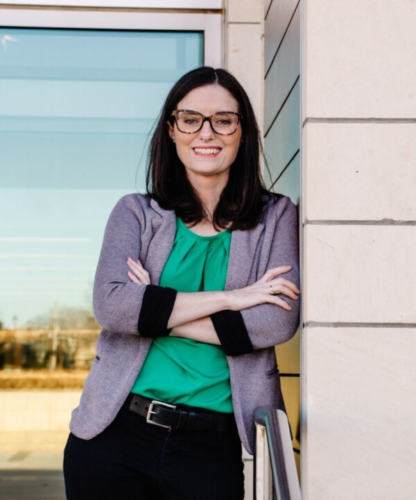 A woman with long dark hair and glasses stands with her arms crossed against a beige wall, wearing a green top and a gray cardigan, smiling confidently.